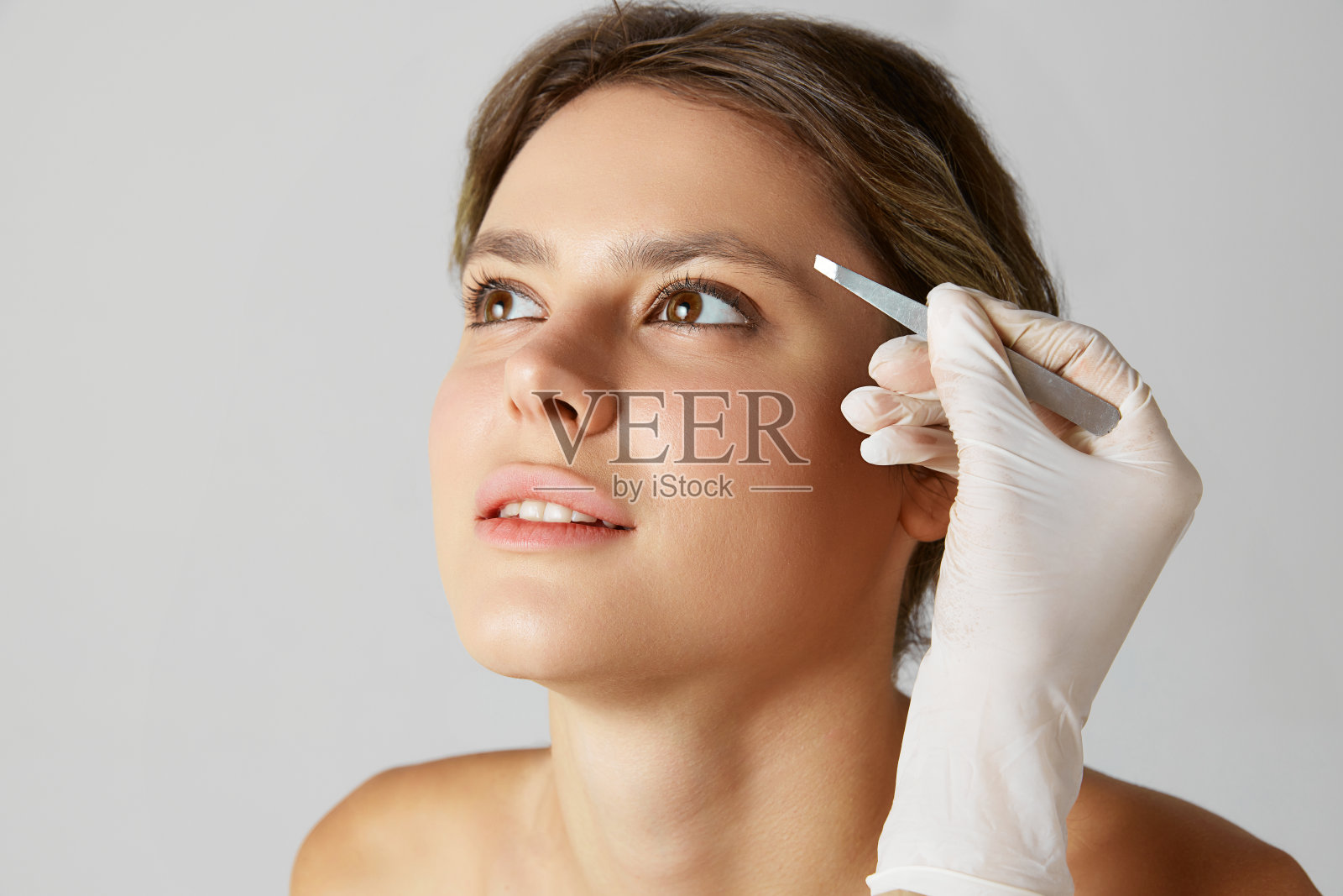 美丽的年轻女子在梳妆后，用镊子在灰色的画室背景下拔眉毛照片摄影图片