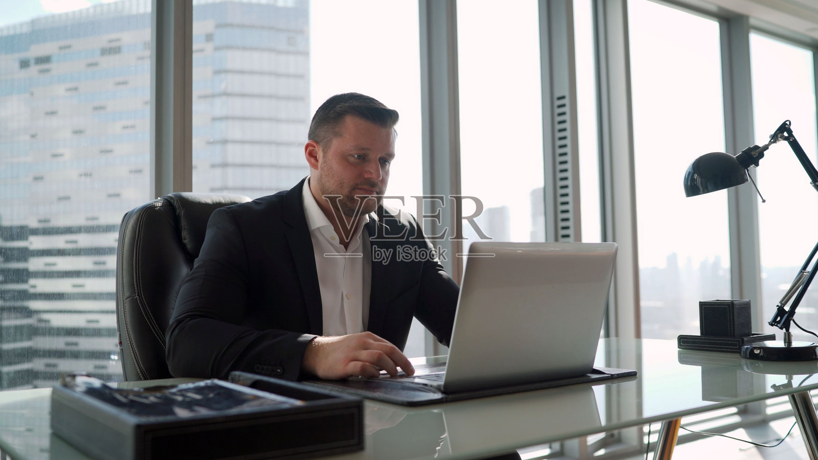 经理在摩天大楼的办公室里用笔记本电脑工作。专注的年轻商人盯着笔记本电脑屏幕，在网上浏览信息或在办公室远程在线工作。一位商人正坐在桌子旁，在一台笔记本电脑上打字照片摄影图片