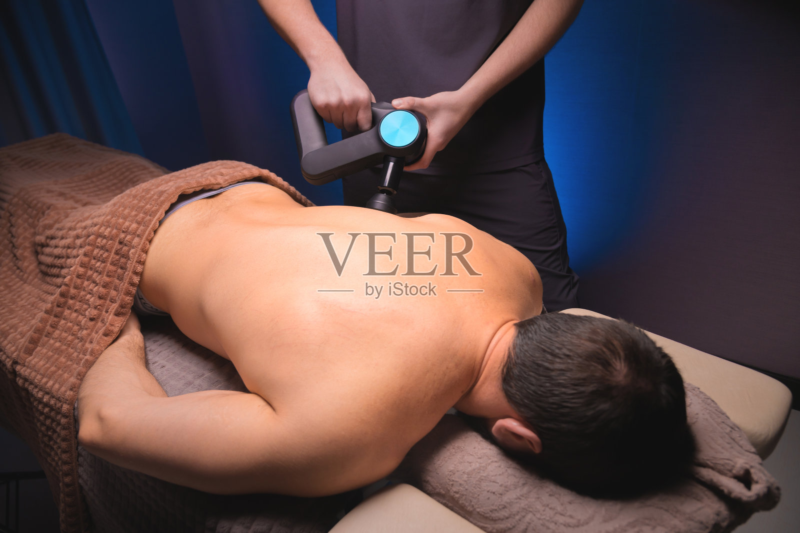 一名男客户躺下后，按摩师手持打击按摩器，对其肩部和上背部进行按摩照片摄影图片