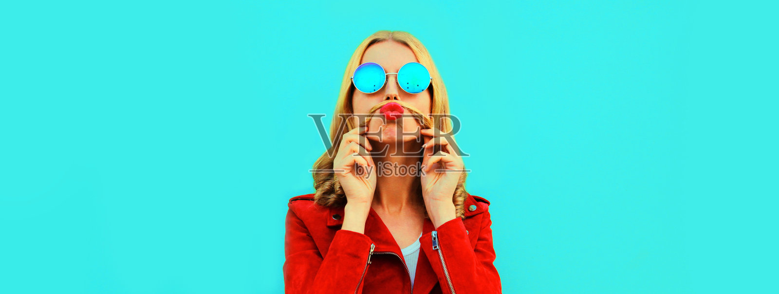 有趣的年轻女子的肖像显示她的胡子，她的头发吹嘴唇与红色的口红发送飞吻戴墨镜在彩色蓝色的背景照片摄影图片