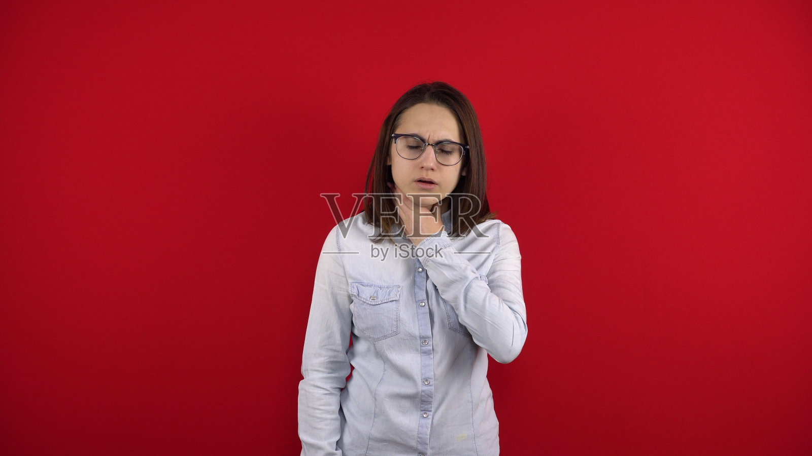 一个戴眼镜的年轻女子喉咙痛，她用手摸了摸他。在红色背景上拍摄。照片摄影图片