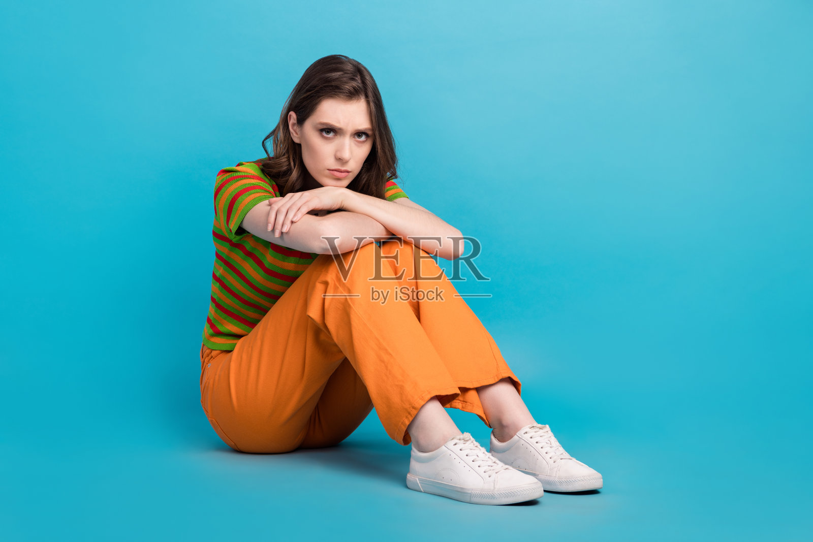 悲伤的女人的全尺寸照片，波浪发型，穿着彩色开衫，坐在地板上，在蓝色的背景下看起来很生气照片摄影图片