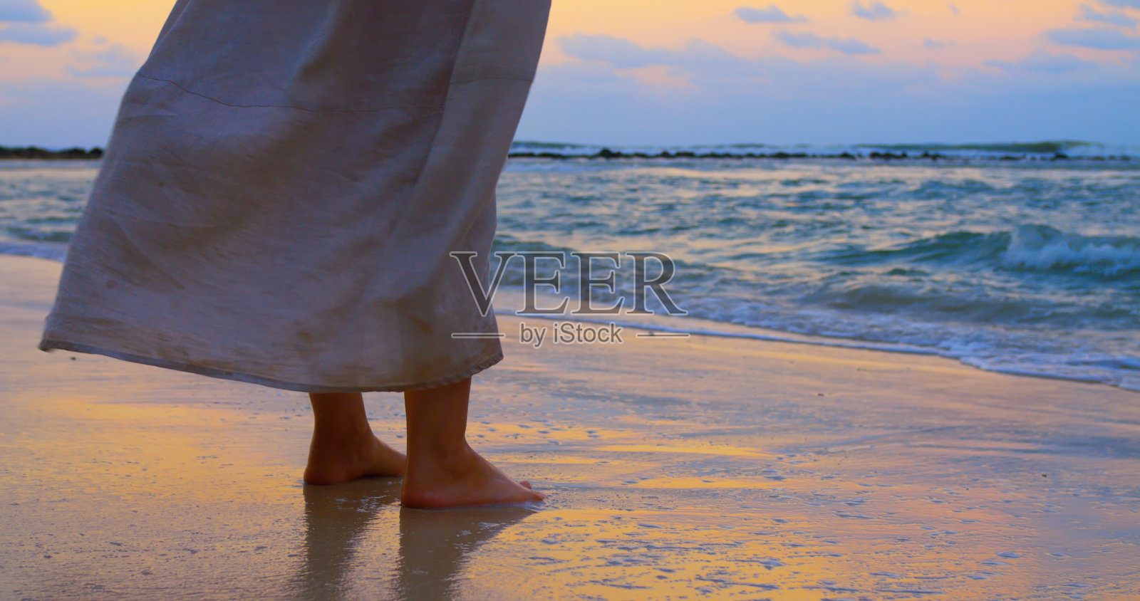 海浪冲刷着脚，风吹着女人的衣服，她站在岸边，欣赏史诗般的日落。平静和凉爽的气氛是让你头脑清醒的最好方式。让波浪在平静的气氛中洗过你的脚照片摄影图片