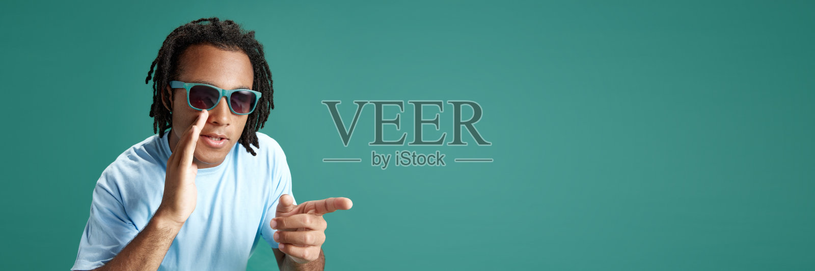 窃窃私语的秘密。戴墨镜的年轻人用手捂着嘴，用手指指着绿色的背景照片摄影图片