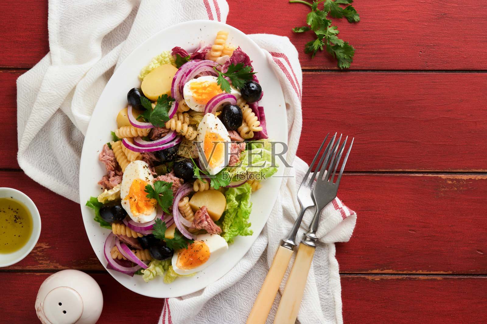 金枪鱼沙拉与意大利面，鸡蛋，土豆，橄榄，红洋葱和酱汁在白色盘子在古老的红色乡村餐桌背景。尼斯沙拉。法国菜。前视图。平的。照片摄影图片