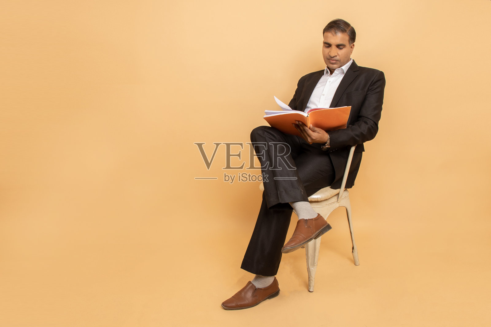 成熟的印度男人穿着黑色西装，坐在米色背景的椅子上看书。副本的空间。照片摄影图片