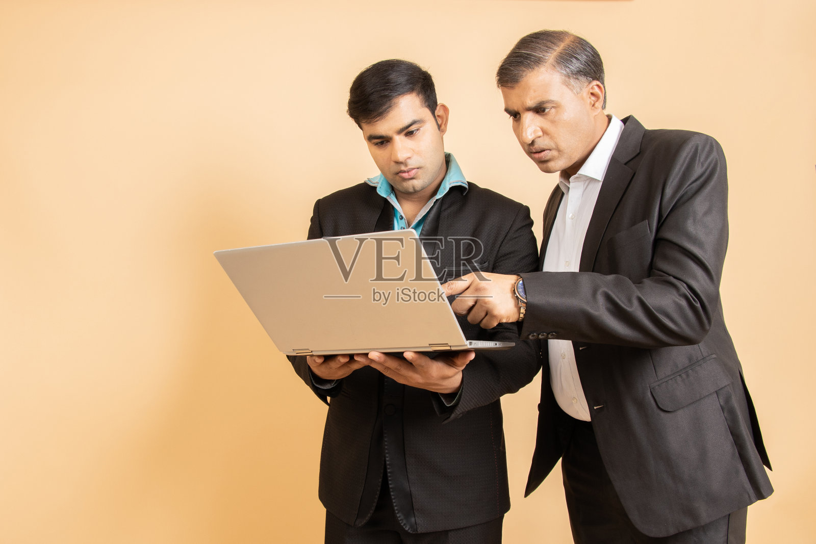 两个穿着黑色正装的印度商人在米色背景上的笔记本电脑上工作。商业和企业理念。照片摄影图片