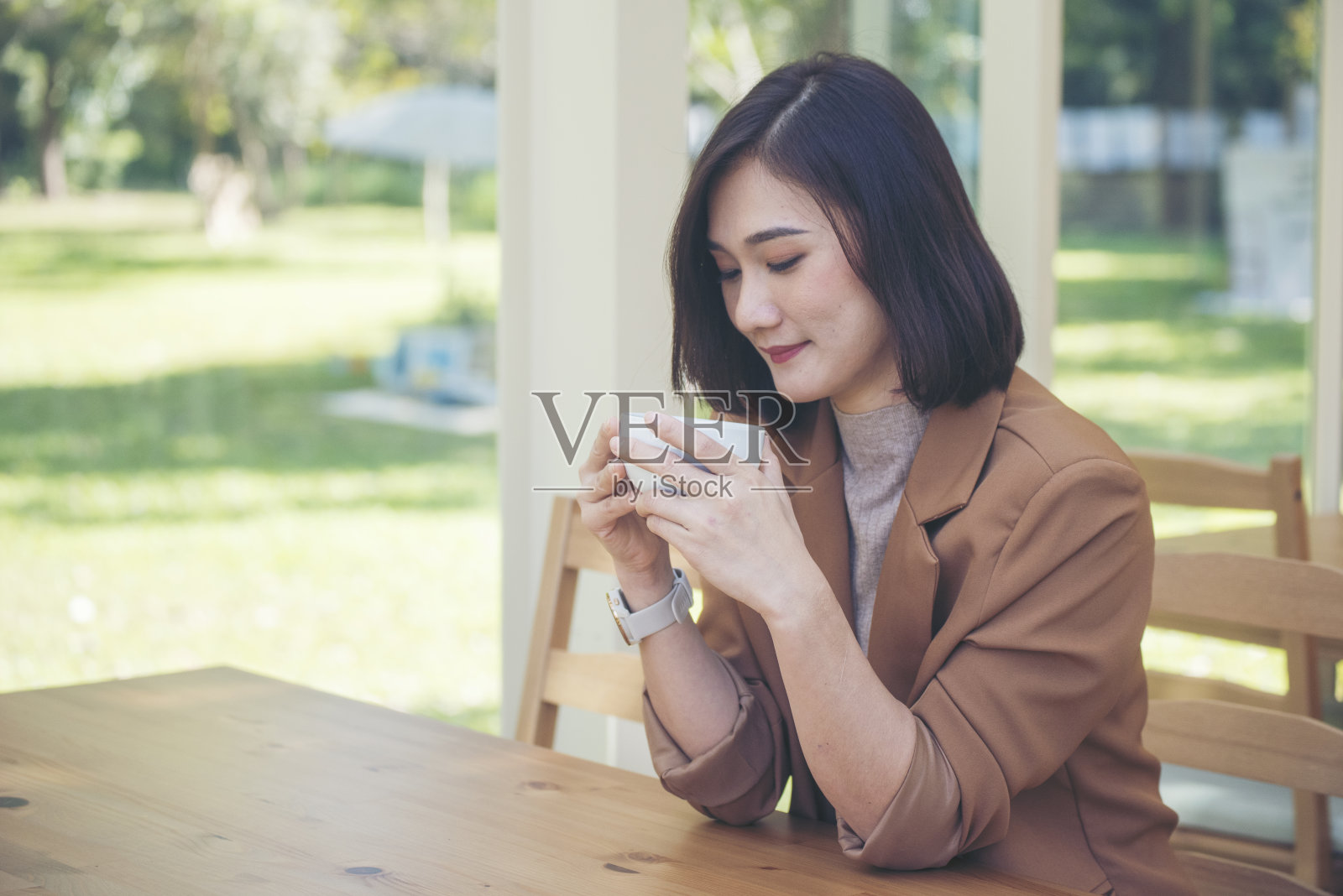 亚洲妇女在绿花园咖啡馆用手捧杯喝黑咖啡。年轻女子笑着脸爱喝咖啡。美女拿着新鲜的杯子在咖啡店喝黑咖啡。咖啡爱好者照片摄影图片
