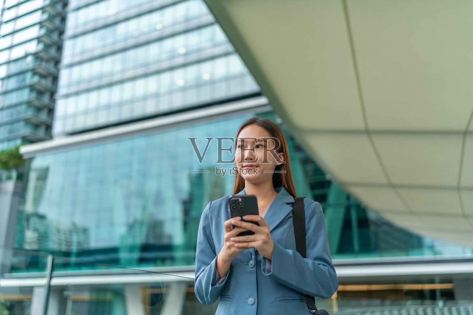 移动中的商业。连接移动。一位年轻的亚洲女商人背着笔记本电脑包，用智能手机上下班，她在中央商务区上班，对面是公司大楼照片摄影图片