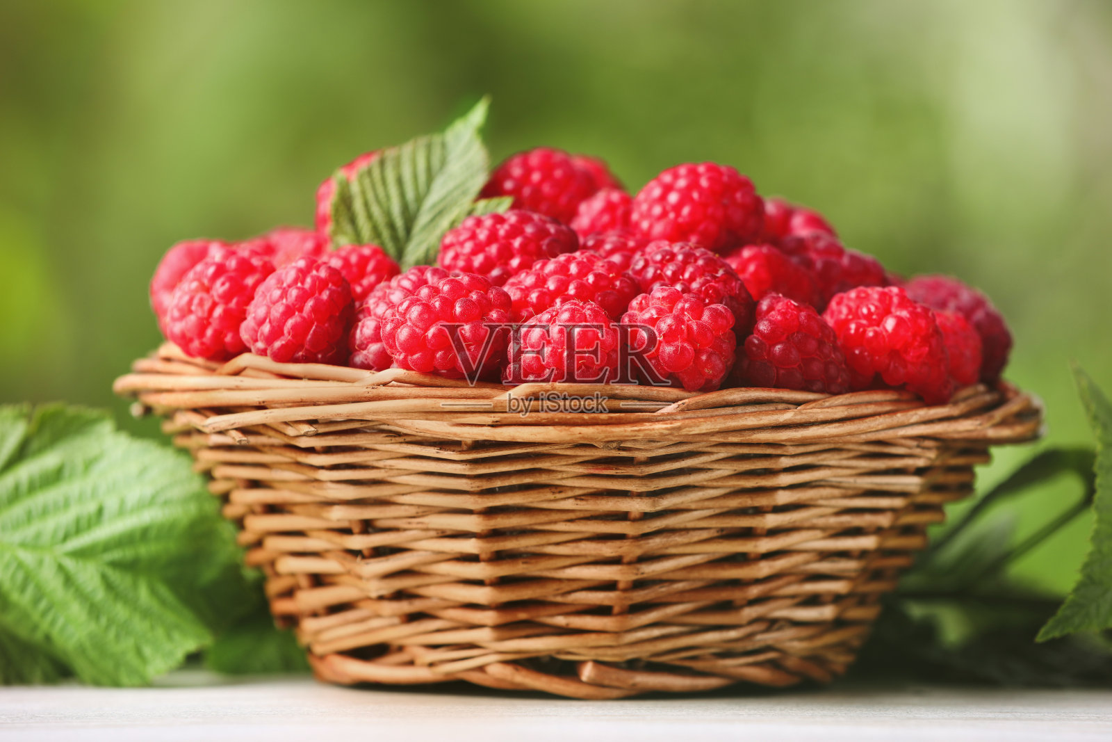 柳条篮子里有美味的成熟树莓和树叶，白色的桌子上有模糊的绿色背景照片摄影图片