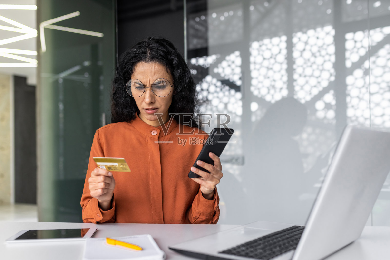 在办公室里沮丧和失望的西班牙裔女商人，拿着银行信用卡和手机，试图进行网上购物时被骗，转账错误照片摄影图片