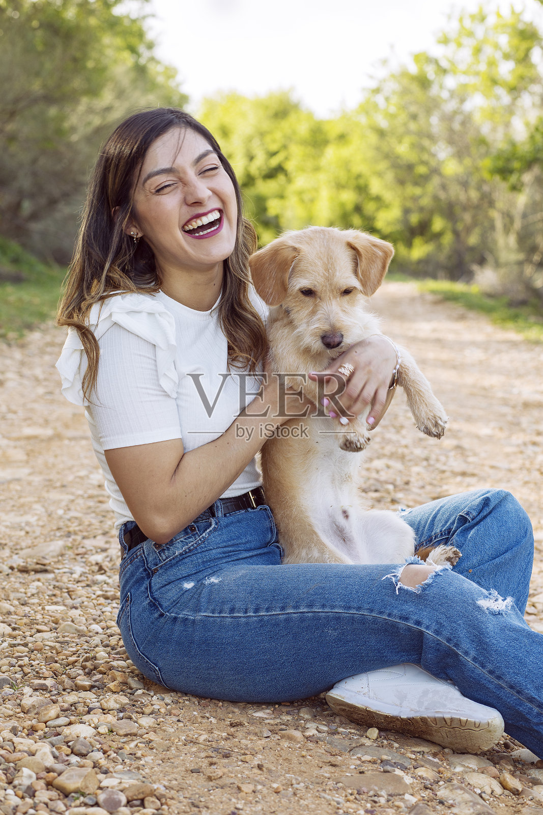 年轻女子笑着，抱着她的狗坐在一条小路上照片摄影图片