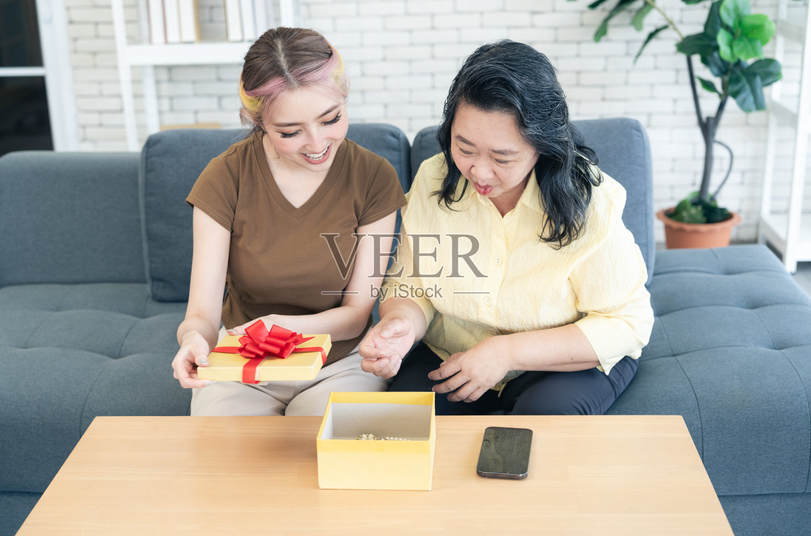 亚洲女儿和母亲坐在客厅的沙发上，激动地打开礼盒或礼物。照片摄影图片