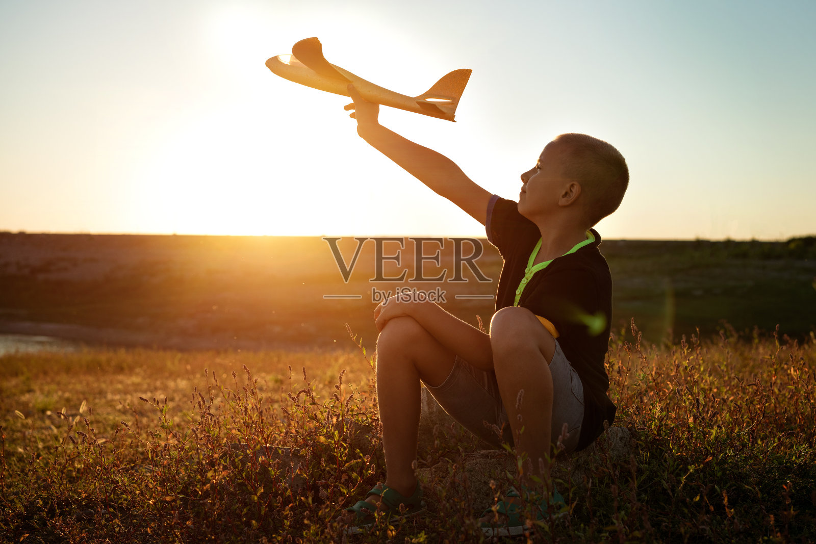 男孩坐在日落，手里拿着一架飞机照片摄影图片