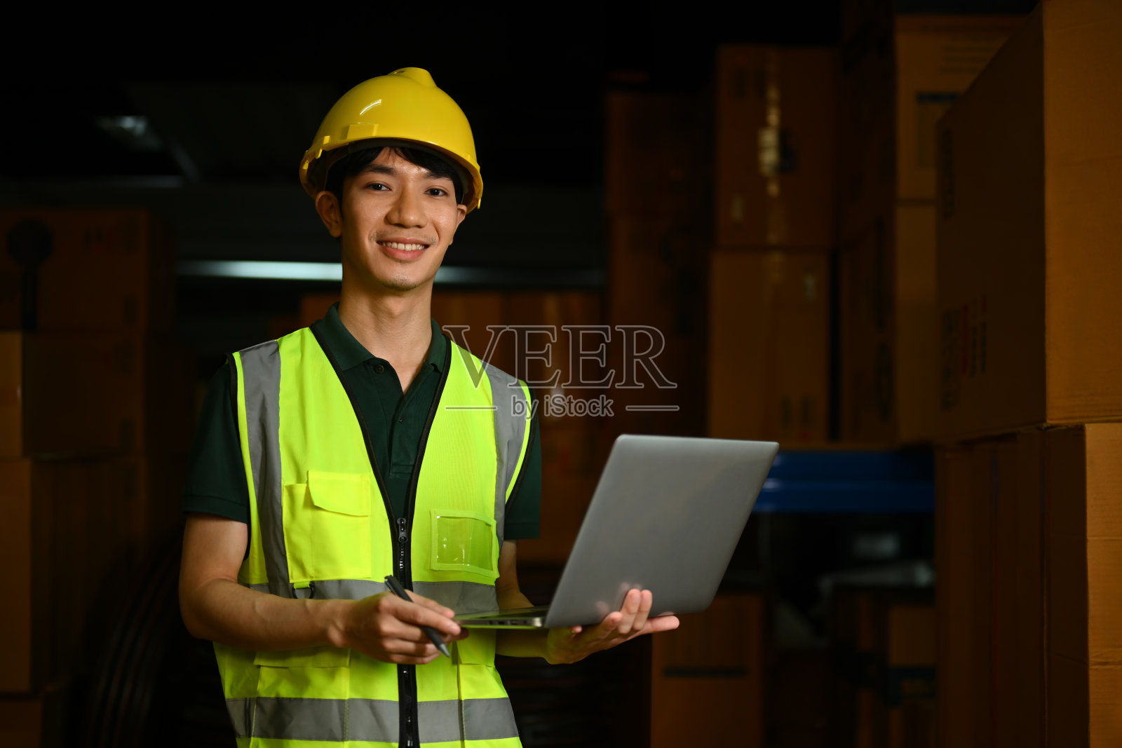 面带微笑的男经理戴着安全帽，穿着反光夹克，站在放着纸板箱的货架之间照片摄影图片