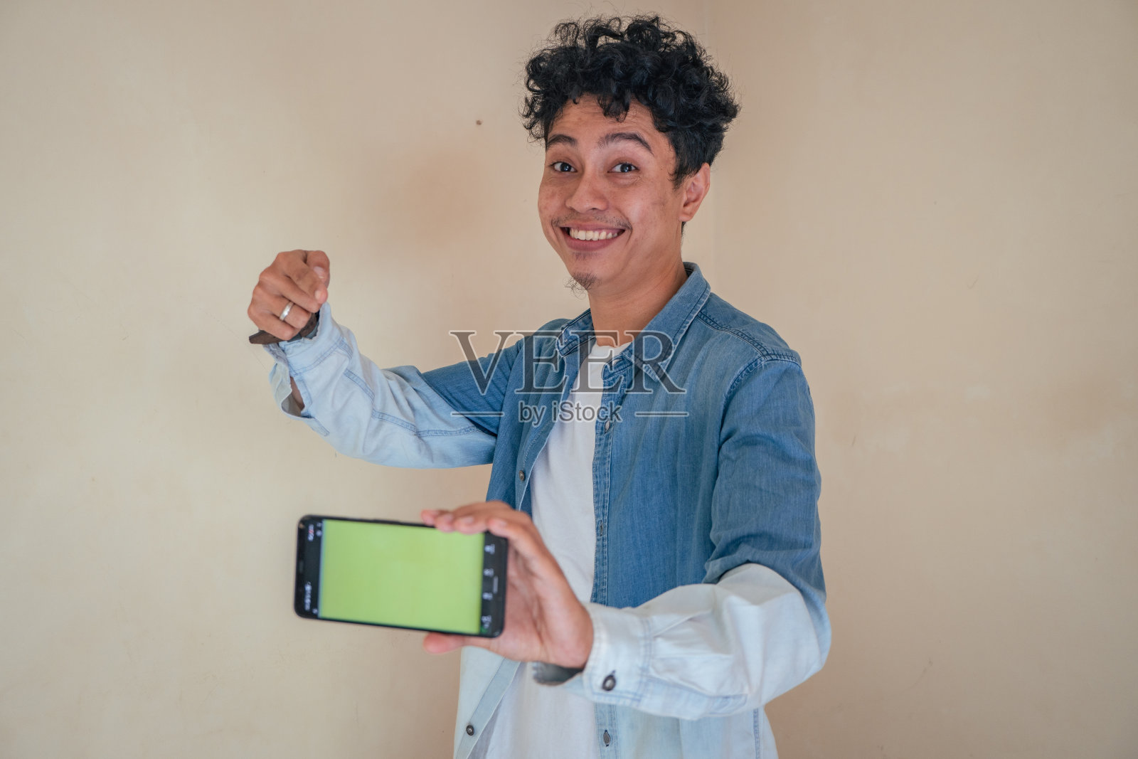 年轻卷曲的男子穿着牛仔衣服，手持绿色屏幕的智能手机。微笑，欢呼和快乐的表情。照片摄影图片