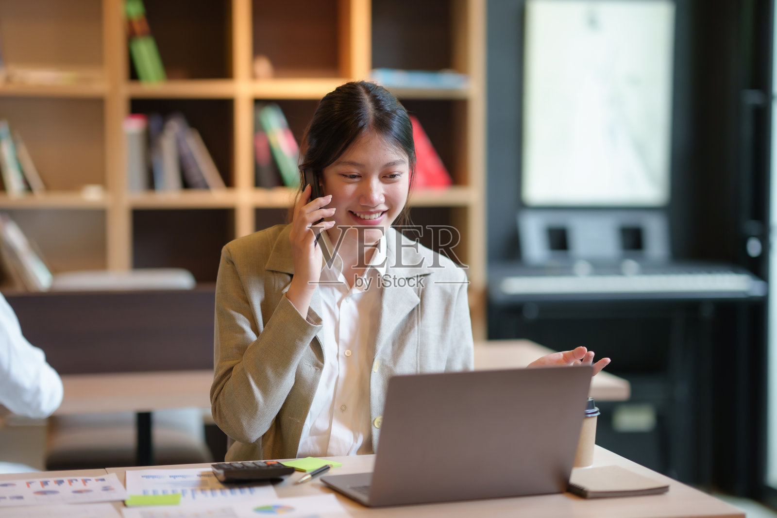 年轻的亚洲女商人坐在她的笔记本电脑前，愉快地与客户在电话中解释细节。照片摄影图片