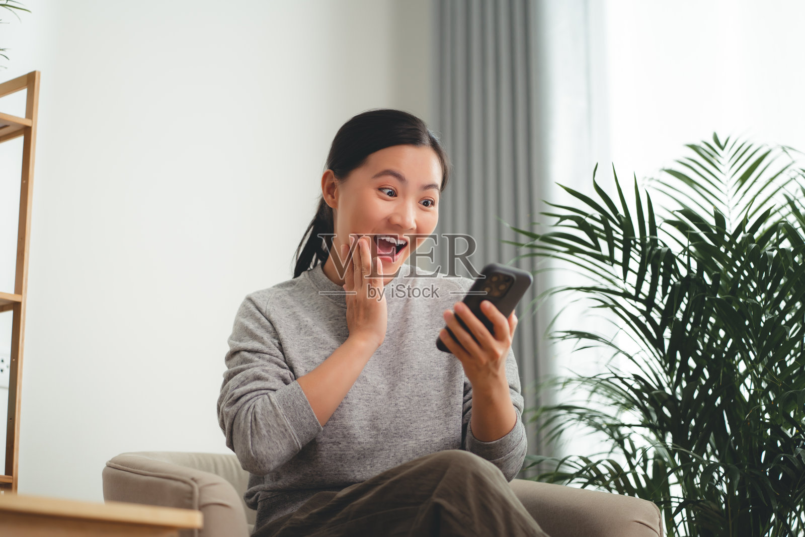 亚洲女性坐在家里的扶手椅上，看着智能手机，开心而惊讶。照片摄影图片