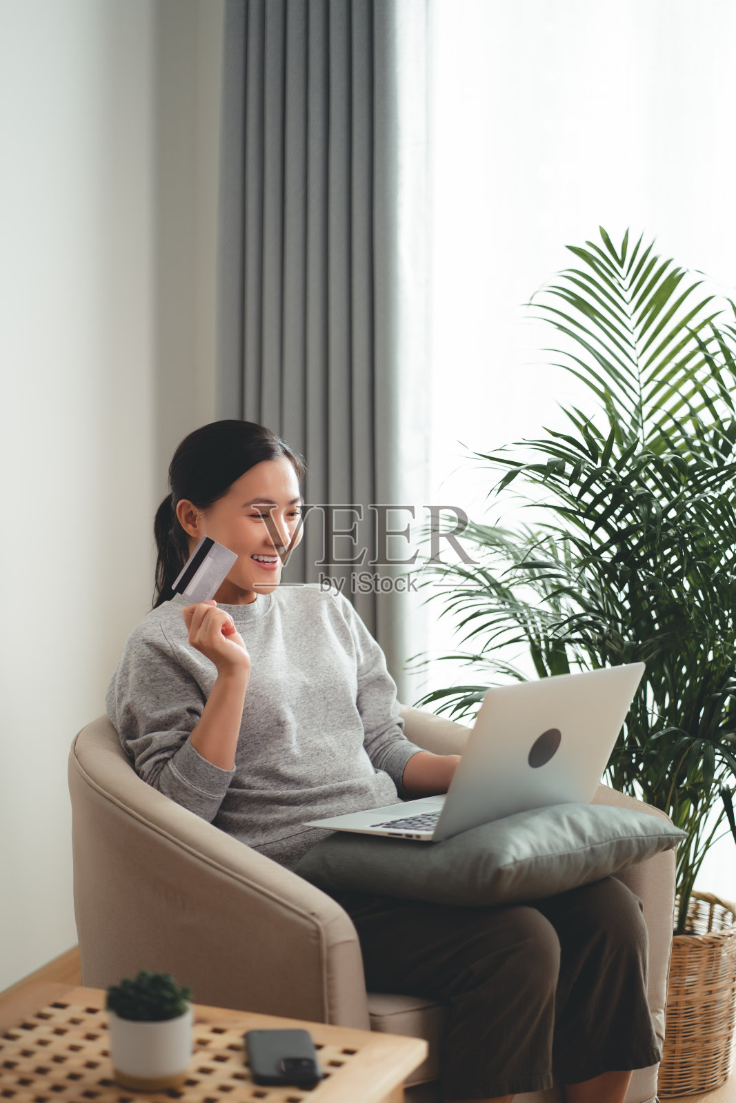 亚洲女人开心地微笑着坐在扶手椅上，用笔记本电脑和信用卡在家网上购物。照片摄影图片