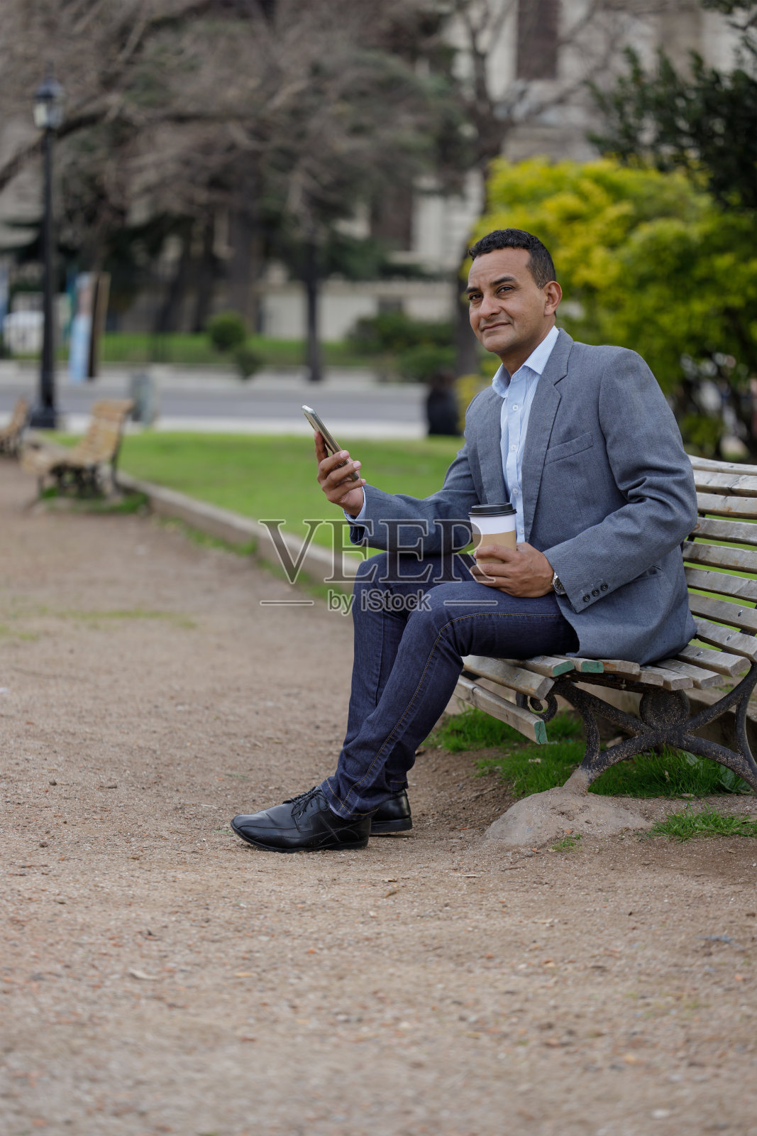 穿着夹克的拉丁裔男子拿着一杯一次性咖啡坐在公园的长椅上。照片摄影图片