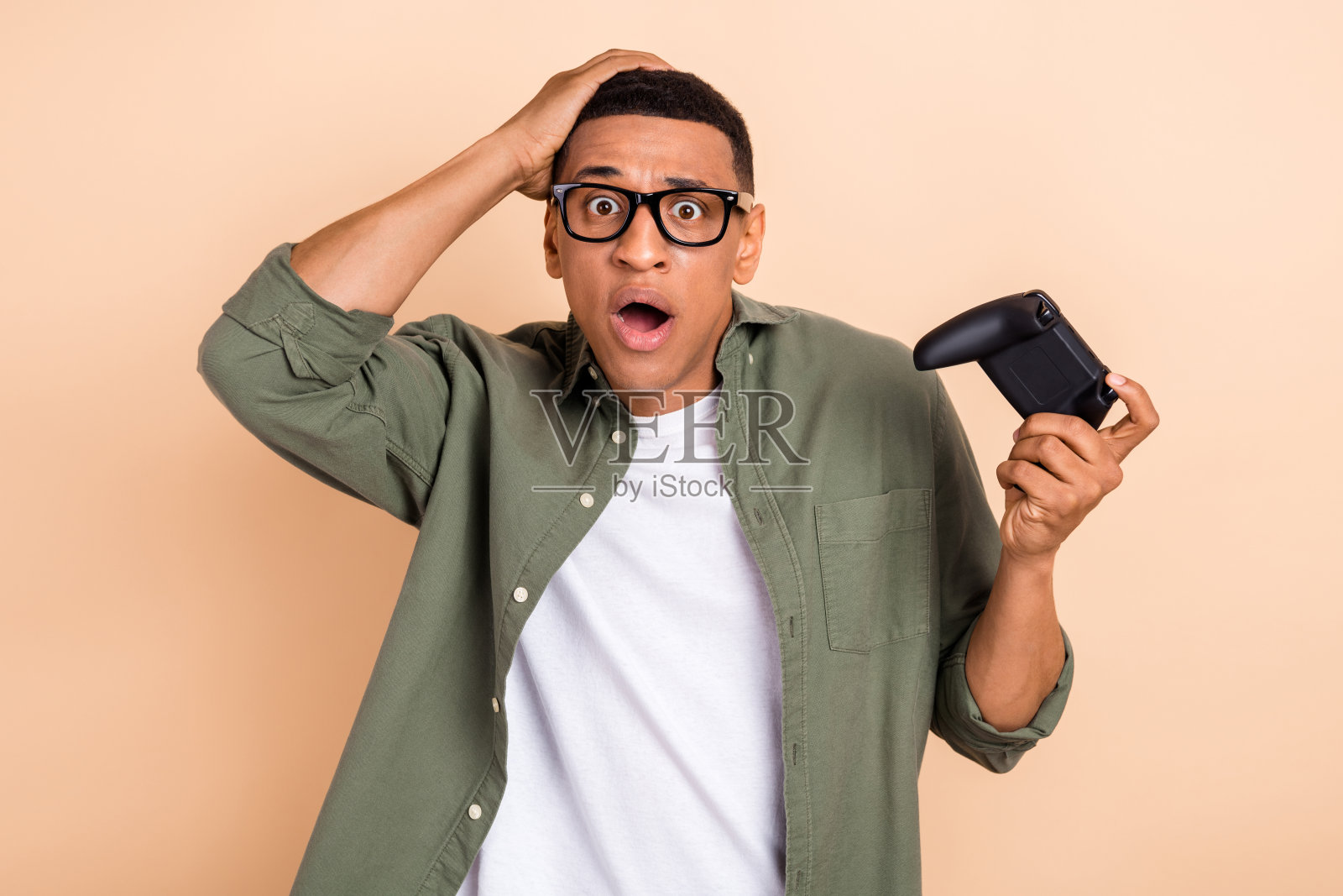 照片中滑稽震惊的男子穿着卡其色衬衫，戴着眼镜，玩着孤立的米色背景游戏照片摄影图片