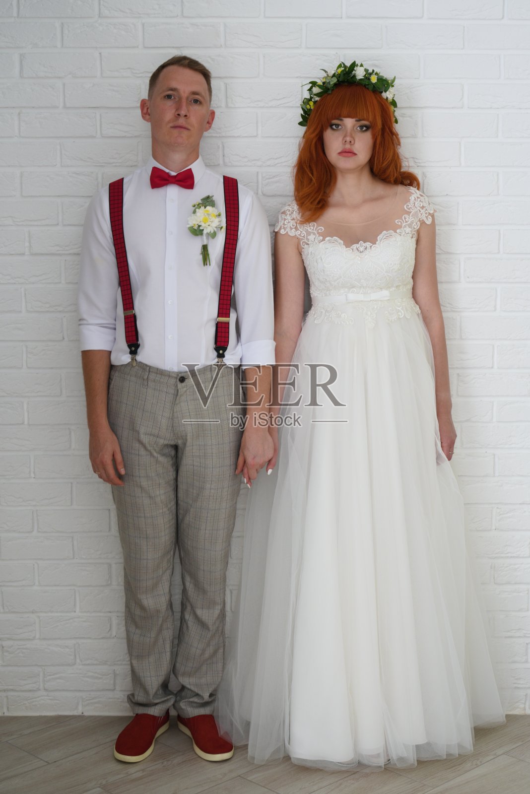 年轻的新娘和新郎面带悲伤的表情，在白色的砖墙旁手牵着手。照片摄影图片