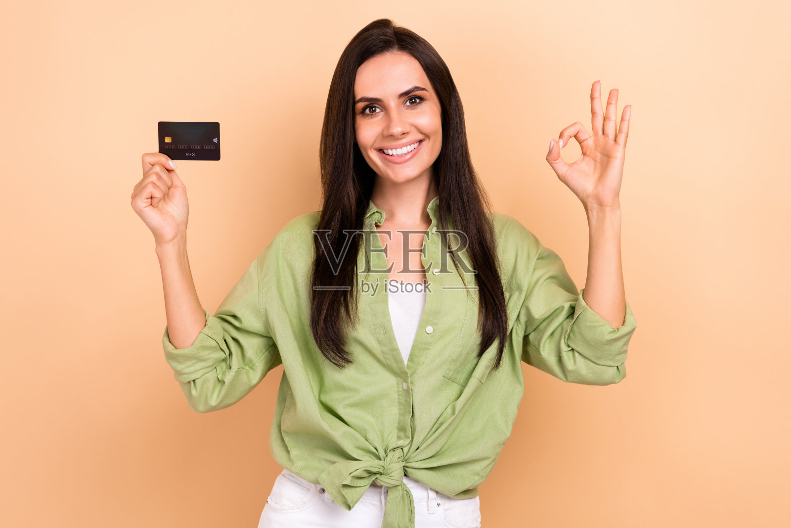 照片中，开朗迷人的女士露齿微笑，拿着借记卡的手指，在米色背景上展示了ok的标志照片摄影图片