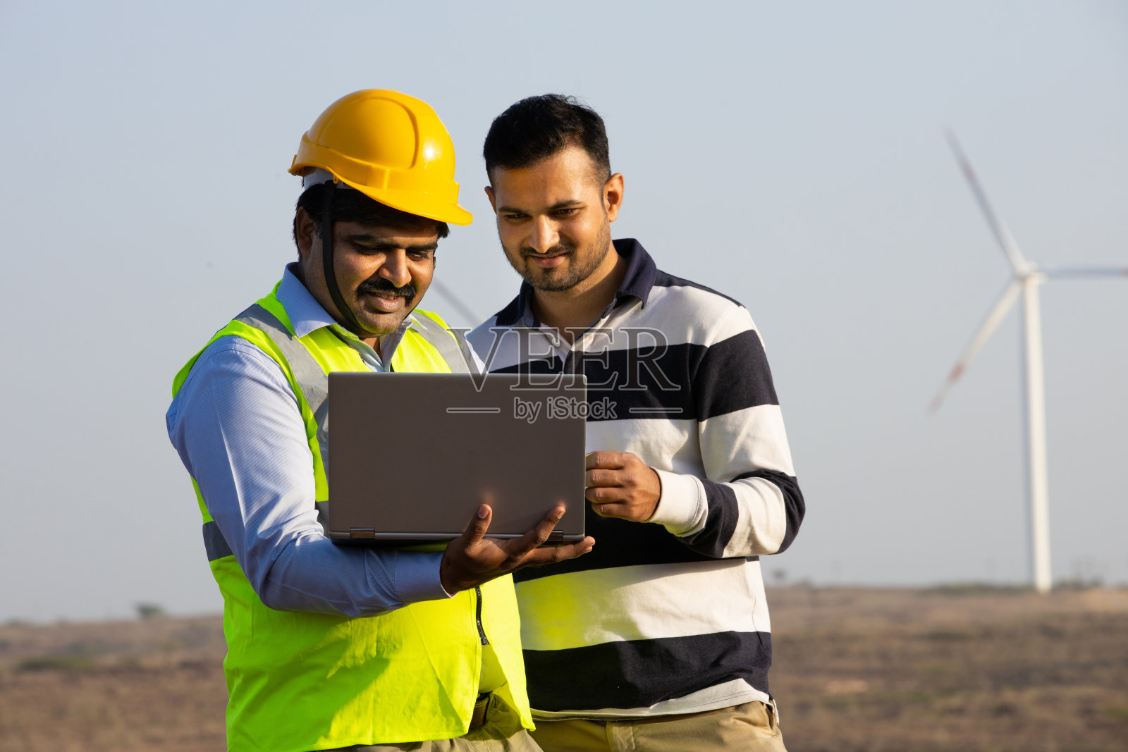 印度工程师使用笔记本电脑，讨论风车农场的运作发电，亚洲人戴着黄色头盔和背心在风力涡轮机农场工作，清洁和绿色能源。照片摄影图片