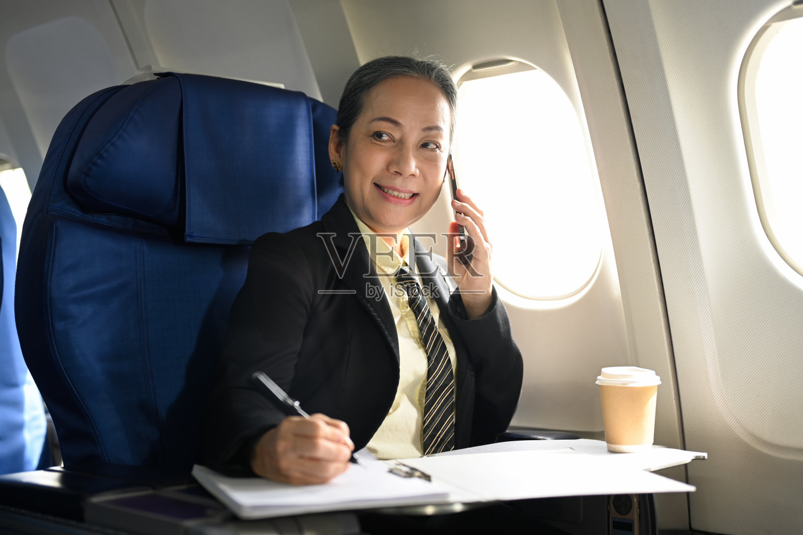 乐观的中年女商人坐在飞机上讲手机。照片摄影图片