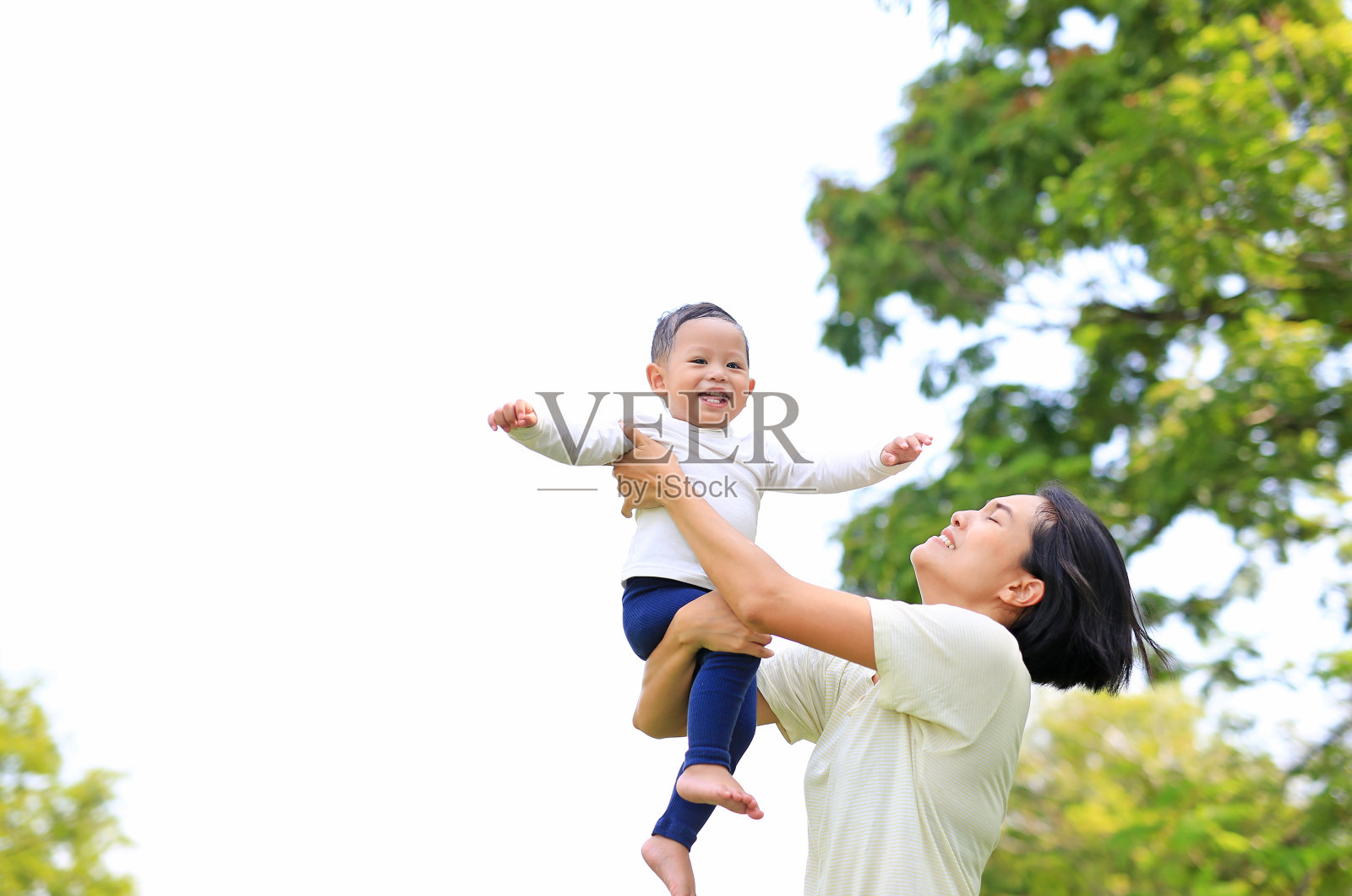 顽皮的亚洲母亲在自然花园里吐出快乐的男婴。母亲在公园里抱着儿子。照片摄影图片