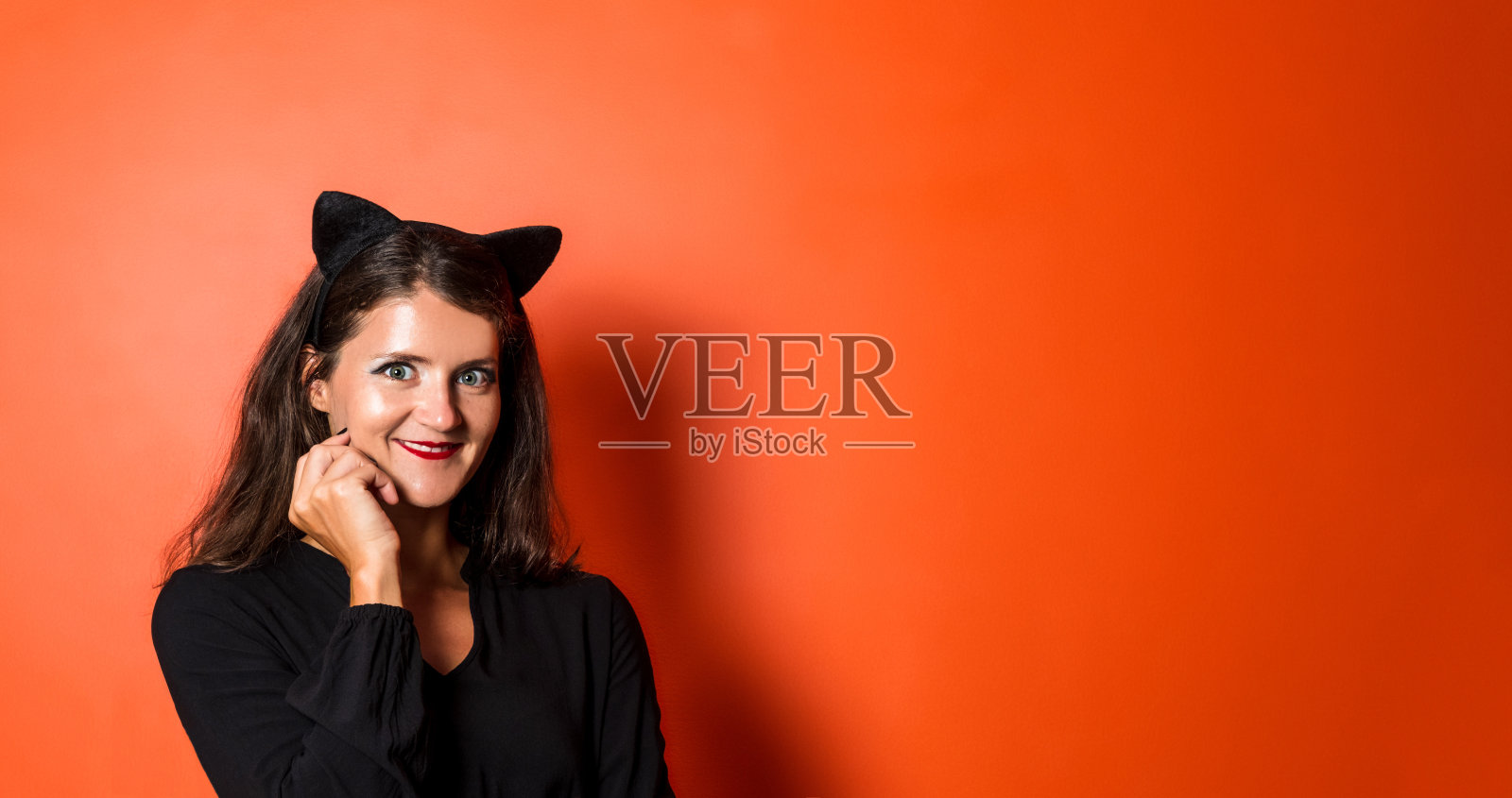 漂亮的年轻女子穿着黑色连衣裙，在橙色的背景上戴着猫耳朵。宽横幅。副本的空间。照片摄影图片