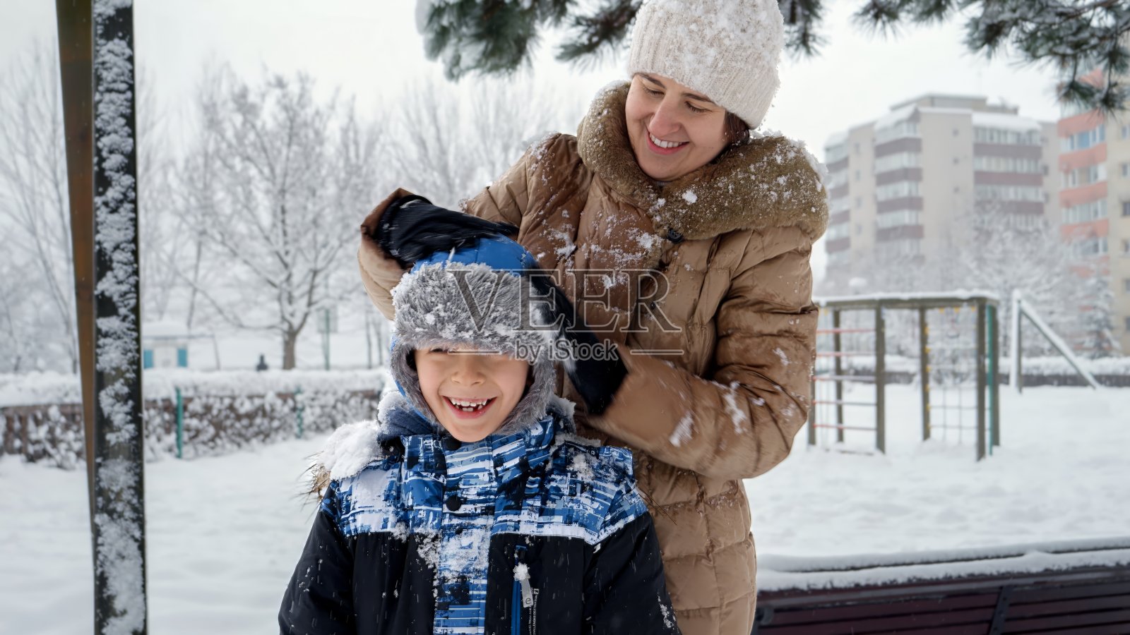 在冬季公园玩耍后，年轻的母亲正在抖掉儿子帽子上的雪。寒假里的欢乐，孩子们在户外玩耍，在雪中活动。照片摄影图片