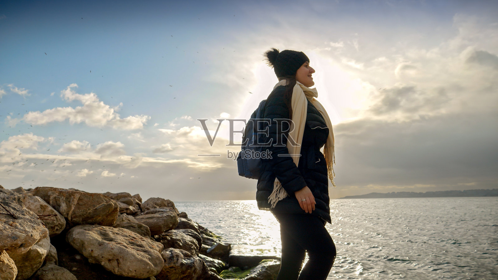 年轻女子背着背包，在日落时分攀爬海边光滑的悬崖。一个冒险，冬季旅游和旅行的概念。照片摄影图片