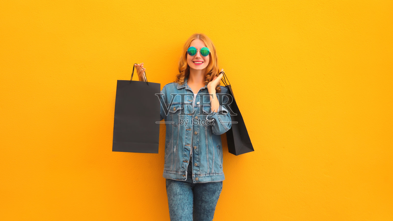 时尚、快乐、微笑的年轻女子在黄色的工作室背景下穿着牛仔夹克，拿着购物袋摆姿势照片摄影图片