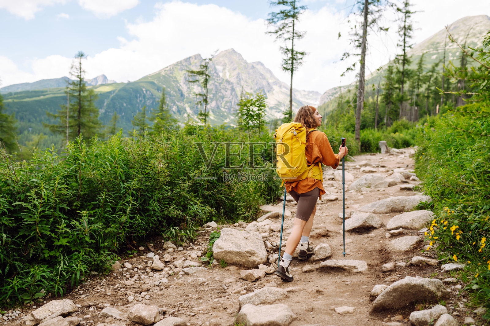 一位时尚的女游客背着黄色背包和登山装备，在山景的背景下沿着登山小径散步。照片摄影图片