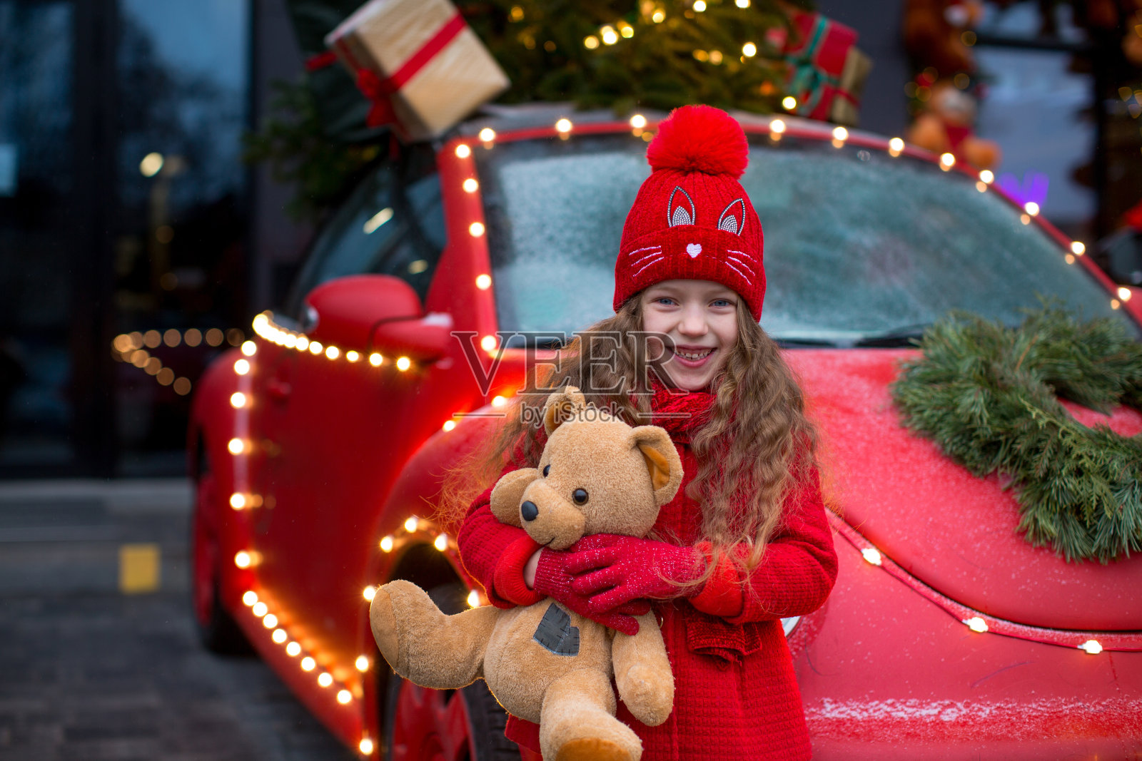 一个快乐的6岁金发女孩穿着红色衣服站在车旁，在圣诞彩灯的背景下拿着一只泰迪熊照片摄影图片