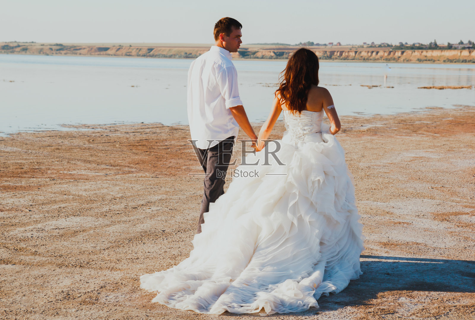 新婚夫妇在海边散步。阳光明媚的夏日照片。新娘披散着头发，穿着露肩裙。海洋浪漫的仪式。海边的爱情故事。沙子，水和地平线。黑色和白色。照片摄影图片