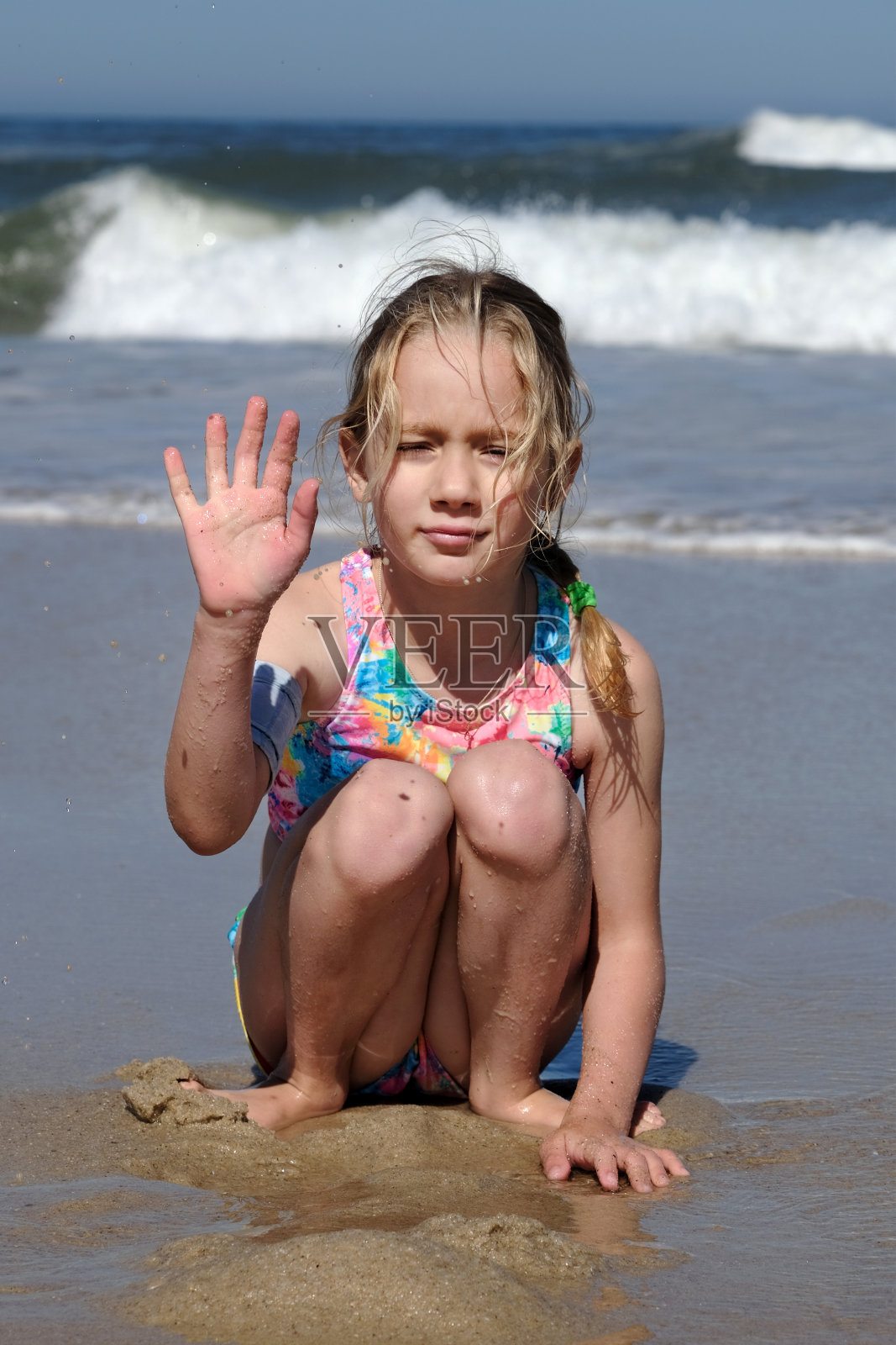 孩子坐在大西洋的背景上，对着镜头挥手。一个七岁的小女孩坐在沙滩上微笑着照片摄影图片
