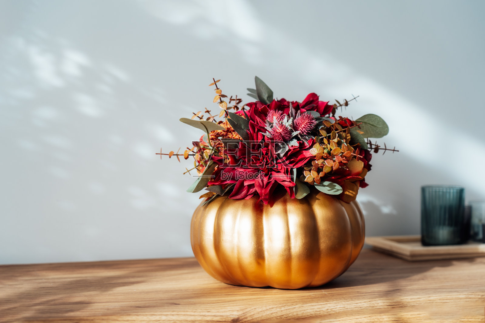 秋天的花束明亮的人造花在一个金色的南瓜花瓶，蜡烛在木制控制台与白色的墙壁与阴影的背景。Hygge家居秋季装饰，营造舒适的家居氛围。照片摄影图片