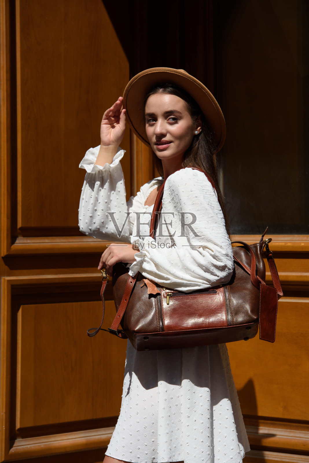 身穿白色浪漫夏装、头戴米色帽子、手拿奢华皮包的黑发女郎照片摄影图片