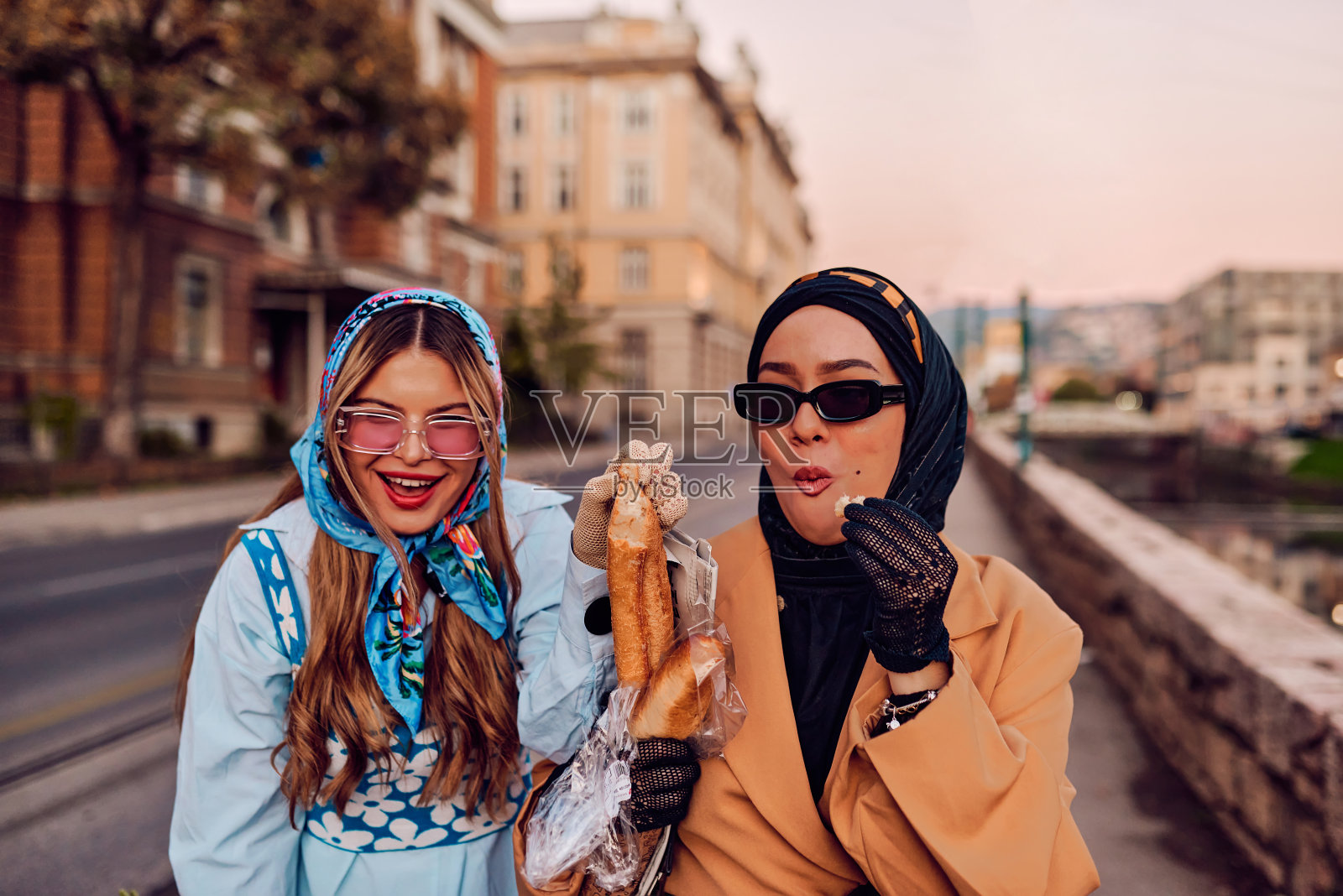 一对妇女，一个戴着头巾，穿着现代而传统的服装，另一个穿着蓝色的裙子和围巾，在日落时分一起穿过城市。一个拿着花束和面包，而另一个拿着一个蓝色的手提包，象征着美丽的fusi照片摄影图片