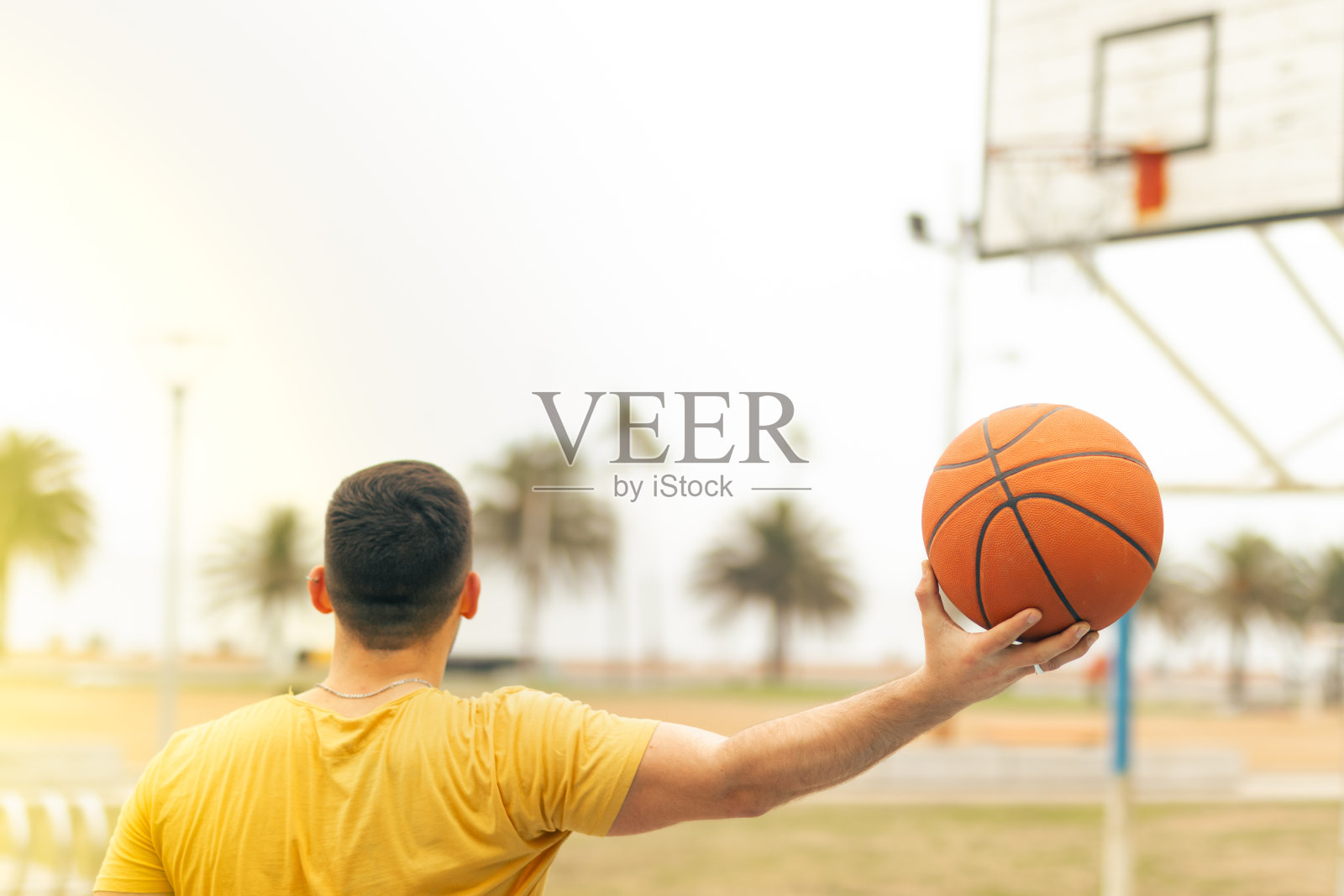 一个年轻人背对着镜头，一只手撑在伸出的手臂上，拿着篮球。模糊的篮球圈背景。胜利的概念。照片摄影图片