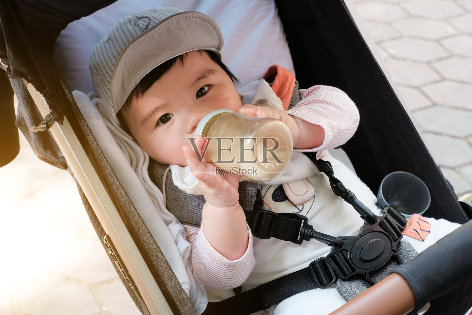 亚洲婴儿戴着帽子，拿着奶瓶，坐在装有安全带的婴儿车里。照片摄影图片