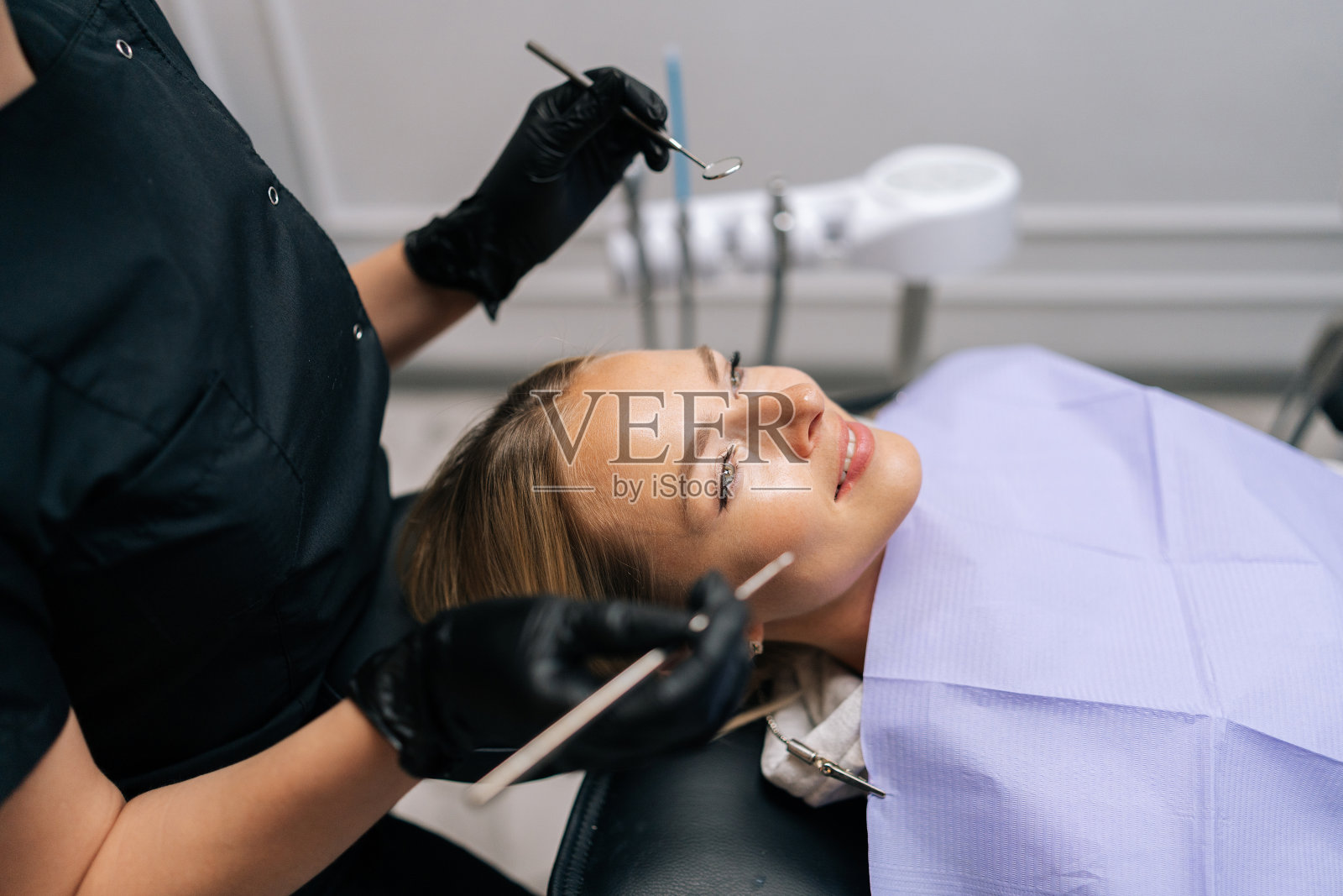 在牙科诊所，无法辨认的女牙医戴着手套检查微笑的年轻女子使用牙科设备仪器的牙齿。照片摄影图片