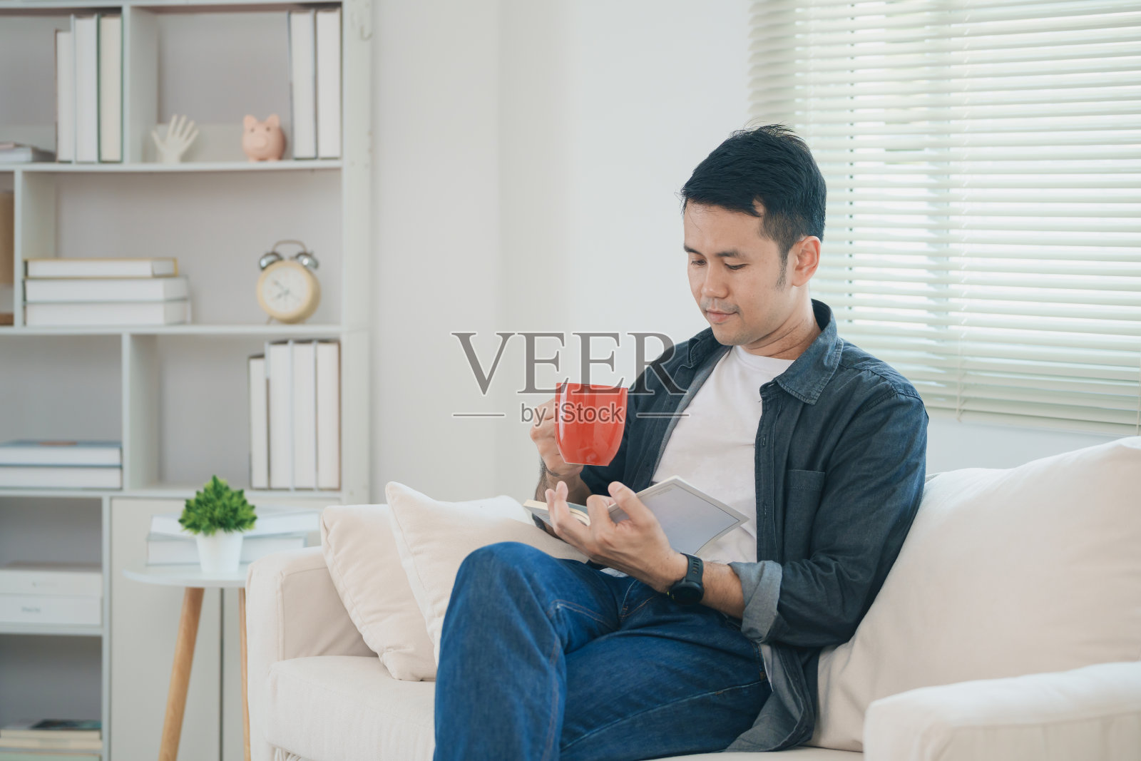 快乐的亚洲男人坐在家里客厅的沙发上看书，喝热咖啡。亚洲男人放松看书，在家里的沙发上微笑。在家活动爱好的概念。照片摄影图片
