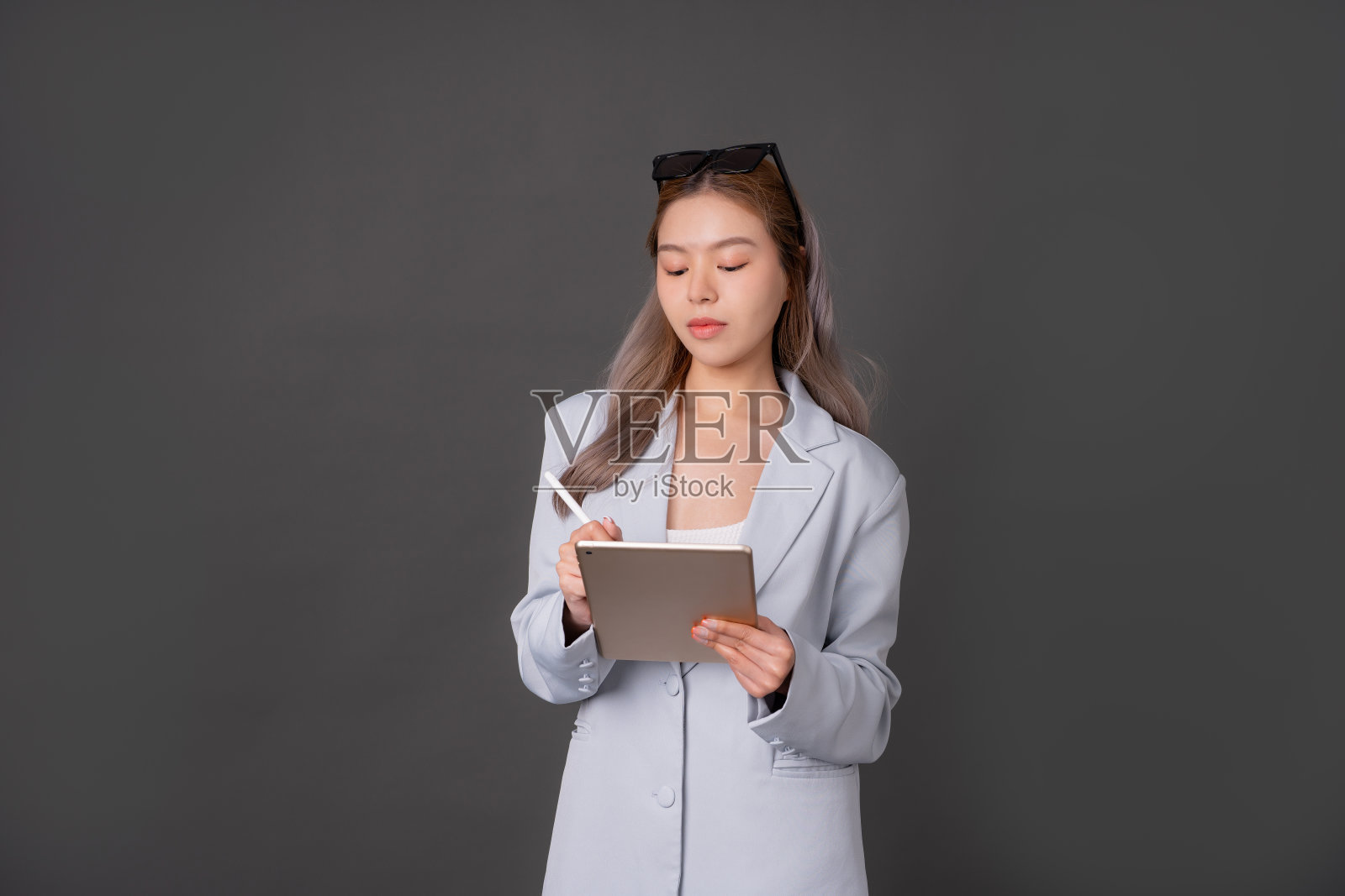 亚洲长发女高管或公司职员，造型干练，戴时尚眼镜，身穿蓝色西装，在灰色背景工作室看平板电脑并用笔工作照片摄影图片