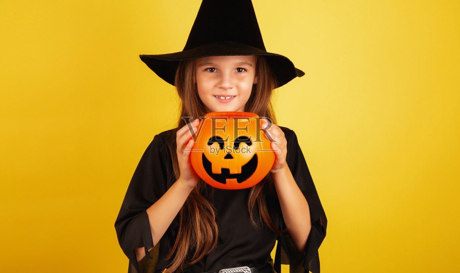 一个长头发的小女孩穿着万圣节的女巫服装微笑着，手里拿着一个黄色背景的南瓜形状的糖果碗。照片摄影图片