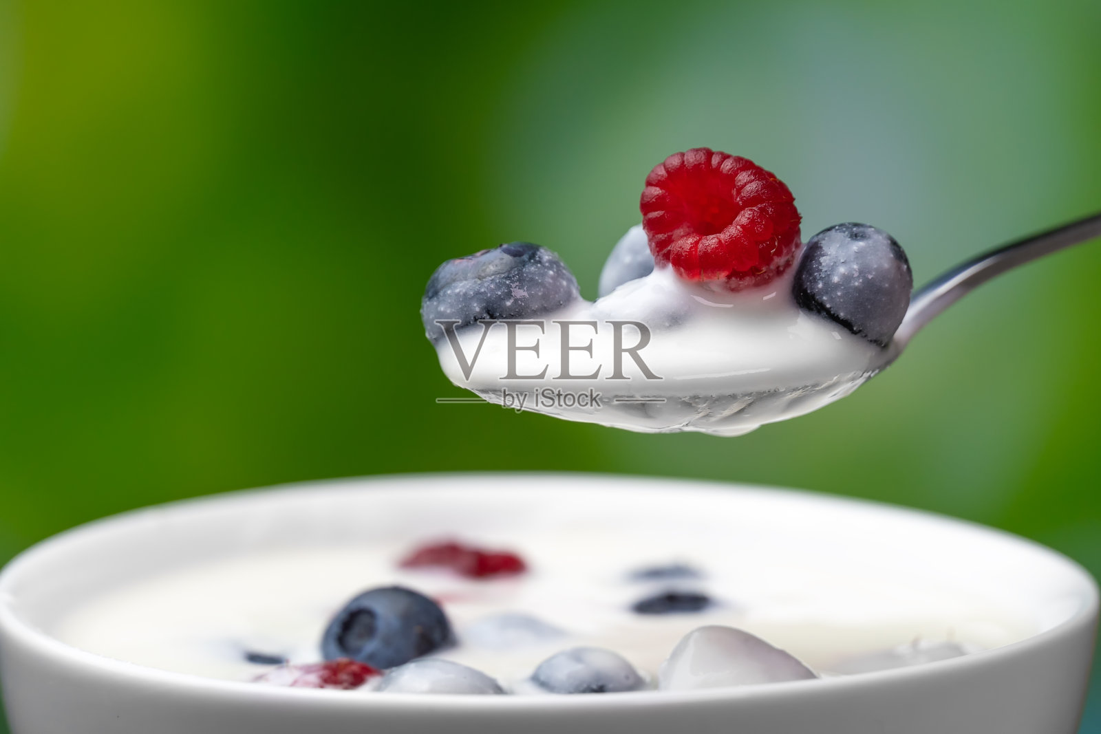 勺子里有希腊酸奶和蓝莓，树莓放在白色的碗里，绿色模糊的背景，微距摄影。由天然食材制成的美味健康早餐照片摄影图片
