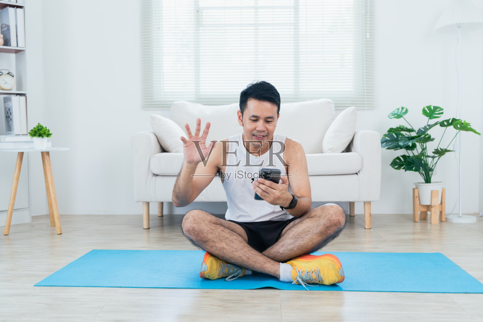 在线课堂培训。快乐的亚洲成熟男子穿着运动服，在客厅室内用手机在垫子上伸展腿，微笑着。快乐的运动员用他的袖子锻炼。在家运动。照片摄影图片