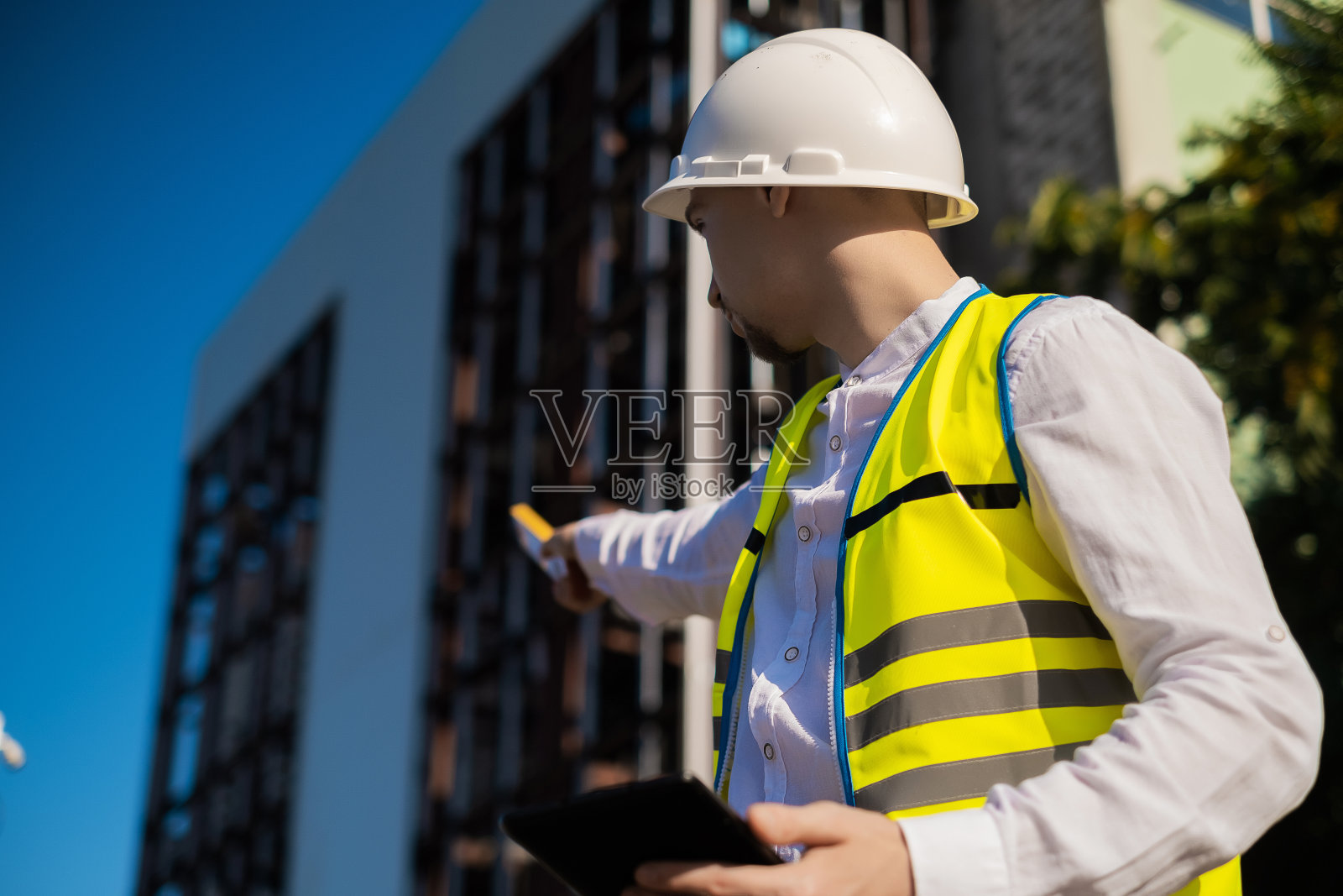 图为在建筑工地工作的建筑师，身穿防护服，头戴安全帽，手拿笔记本电脑，指着屏幕左侧照片摄影图片